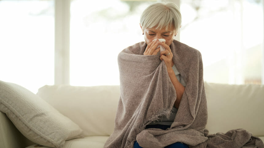 Woman on couch blowing her nose with blanket wrapped around her