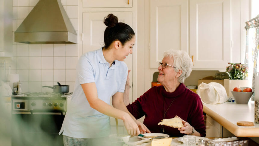 Smiling retired elderly woman talking with female caregiver in kitchen at nursing home
