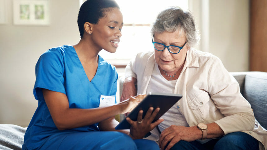 A senior woman using a digital tablet sits next to a nurse on the sofa at home