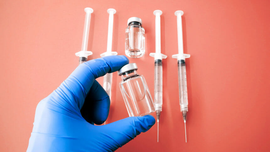 Close-up of gloved hand holding medical vial with flat lay of syringes and medical vial on orange background