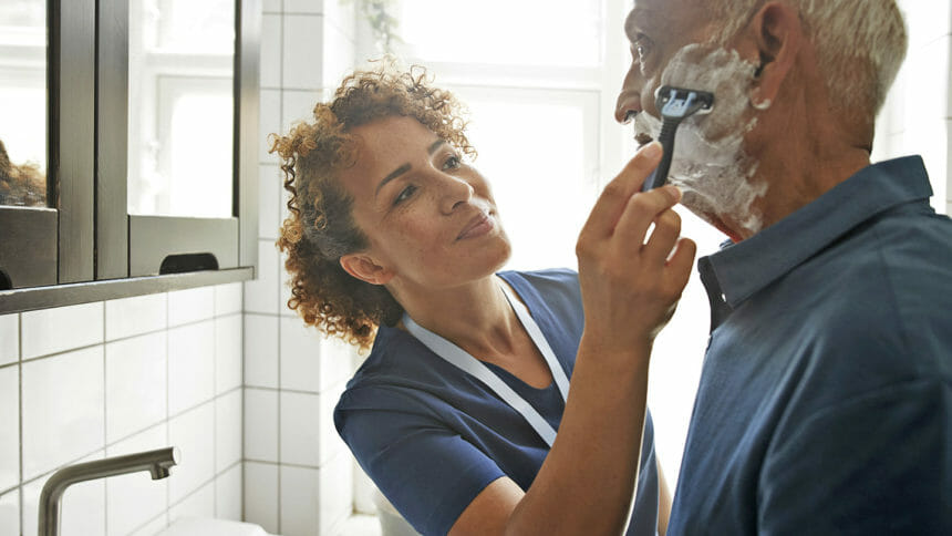 Female healthcare worker assisting senior man in shaving at bathroom