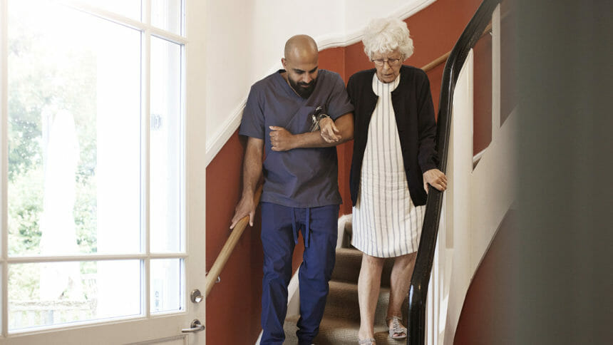 Male caregiver arm in arm with senior woman hand while moving down on steps at home