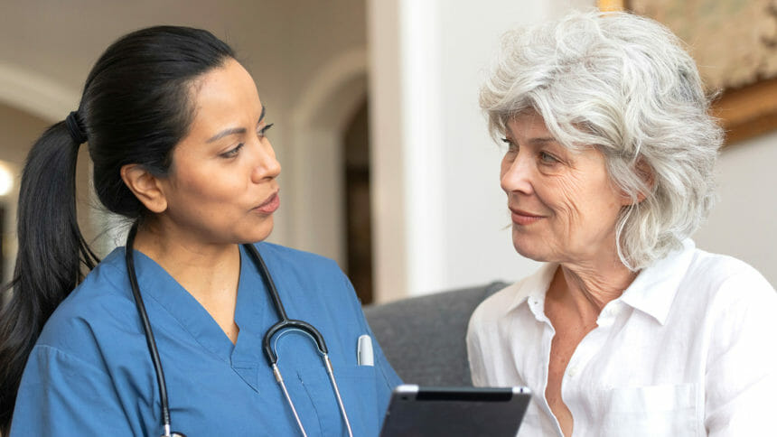A female doctor of Latin descent consults a senior woman in a nursing home. The doctor is using a tablet to show her results from her most recent test.