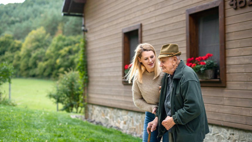 family caregiver, or unpaid caregiver, helps an older adult walk outside in front of a house.
