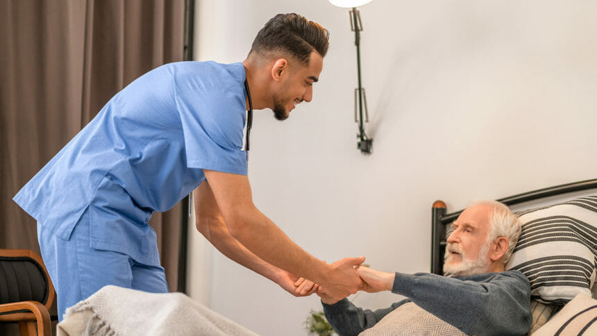 Experienced in-home male nurse in uniform helping an aged patient to rise from the bed