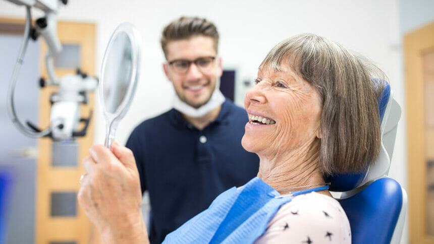 Smiling elderly patient checking her teeth into a hand mirror after her dental treatment.
