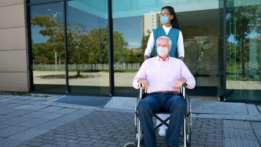 Nurse pushing a senior Latin American man on a wheelchair leaving the hospital and wearing facemasks during the COVID-19 pandemic