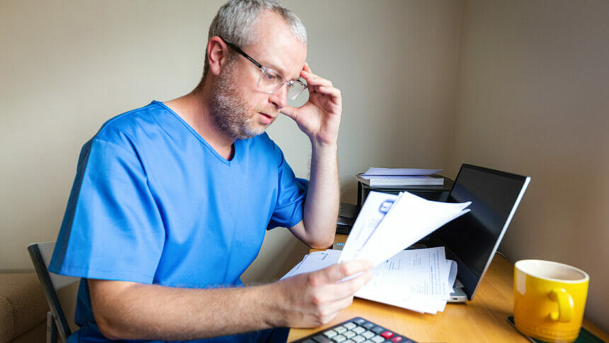 A nurse sits at his desk looking over bills with an expression of financial stress.