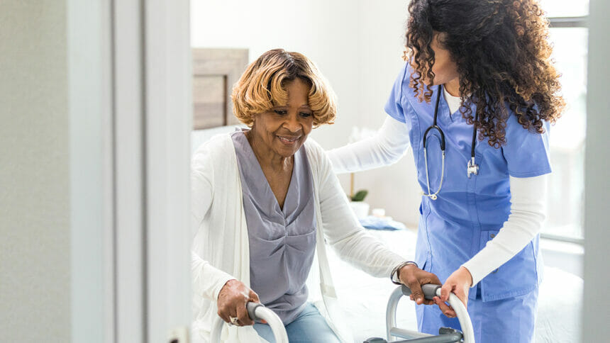 A caring young adult female nurse helps a senior woman stand up. The senior woman is holding on to a walker as she prepares to stand.