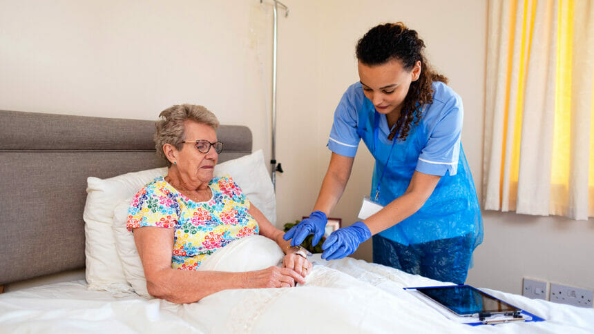 Medium view of a community nurse visiting a senior woman who is lying in her bed at her home in Northumberland, England. The nurse is wearing her scrubs and attaching an IV drip to her patients hand.