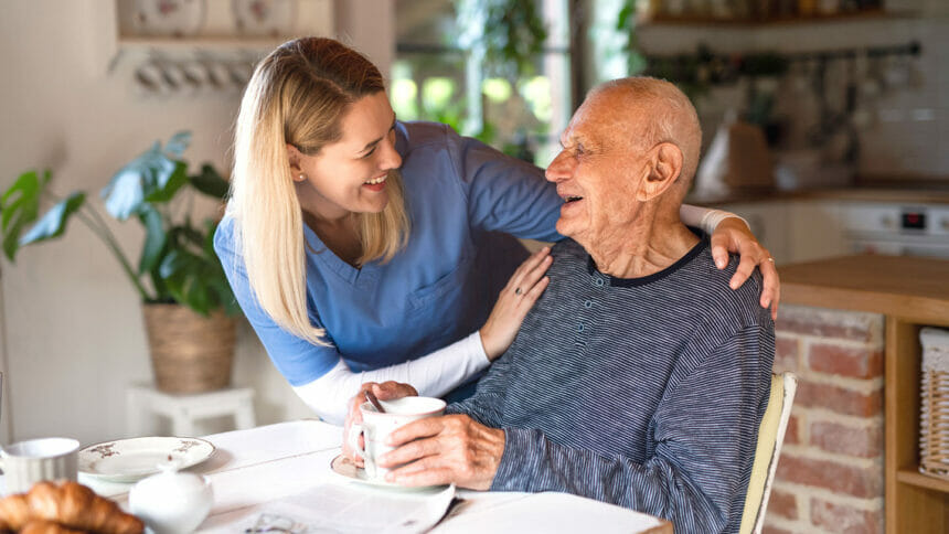 Happy young female caregiver or healthcare worker visiting senior man indoors at home, talking and smiling.