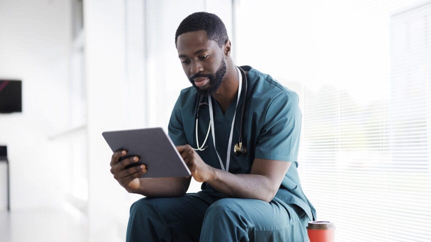 Male doctor sitting in hospital hallway, looking at digital tablet