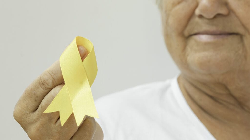 Senior woman is holding yellow awareness ribbon in front of white background. Some issues for what the yellow awareness ribbon stands for are Bone Cancer,Adoptive Parents,Obesity,Missing Children,Missing Persons,Refugees Welcome,Prisoners of War and Suicide Prevention